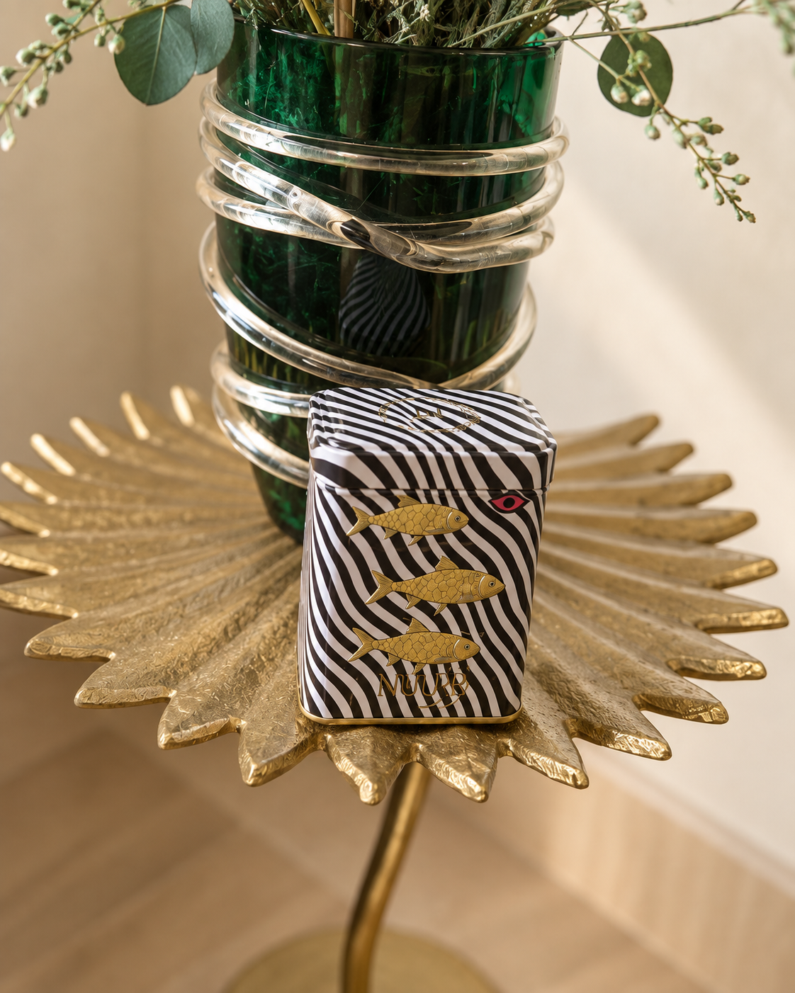 NUUR fish art tin beside an emerald vase on a golden leaf table